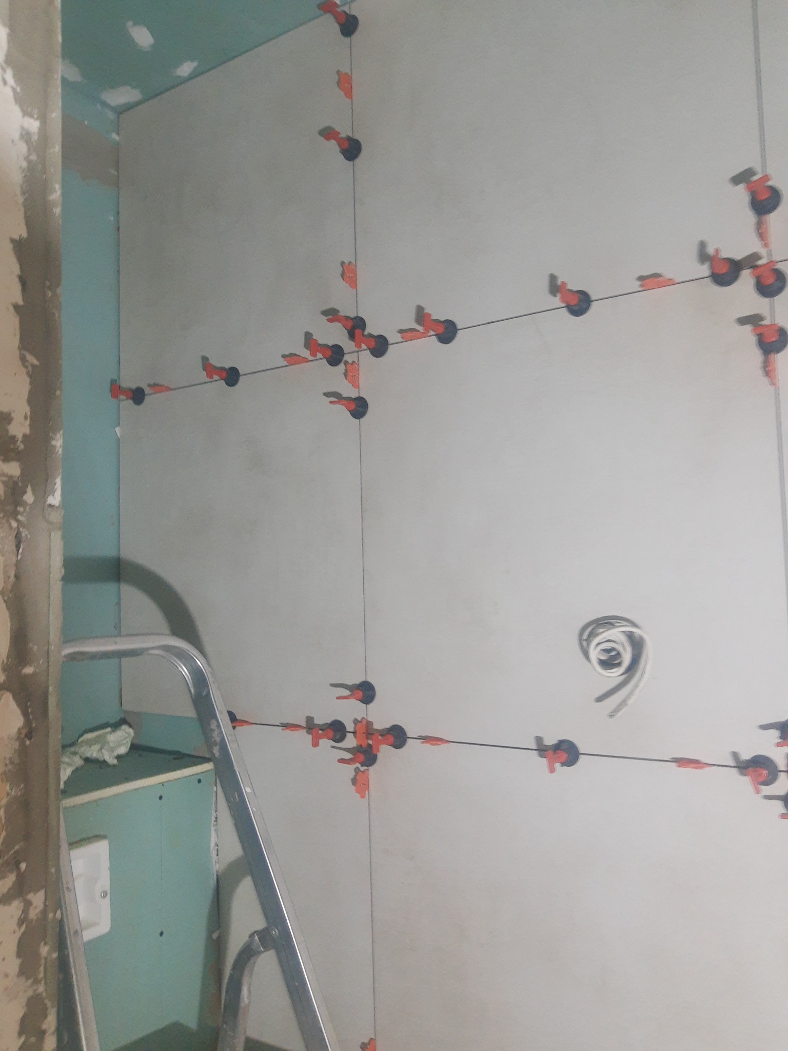 Ściana w trakcie układania płytek, widoczne systemy poziomowania płytek (klipsy i kliny), fragment drabiny i surowa ściana obok, przygotowywana instalacja elektryczna.