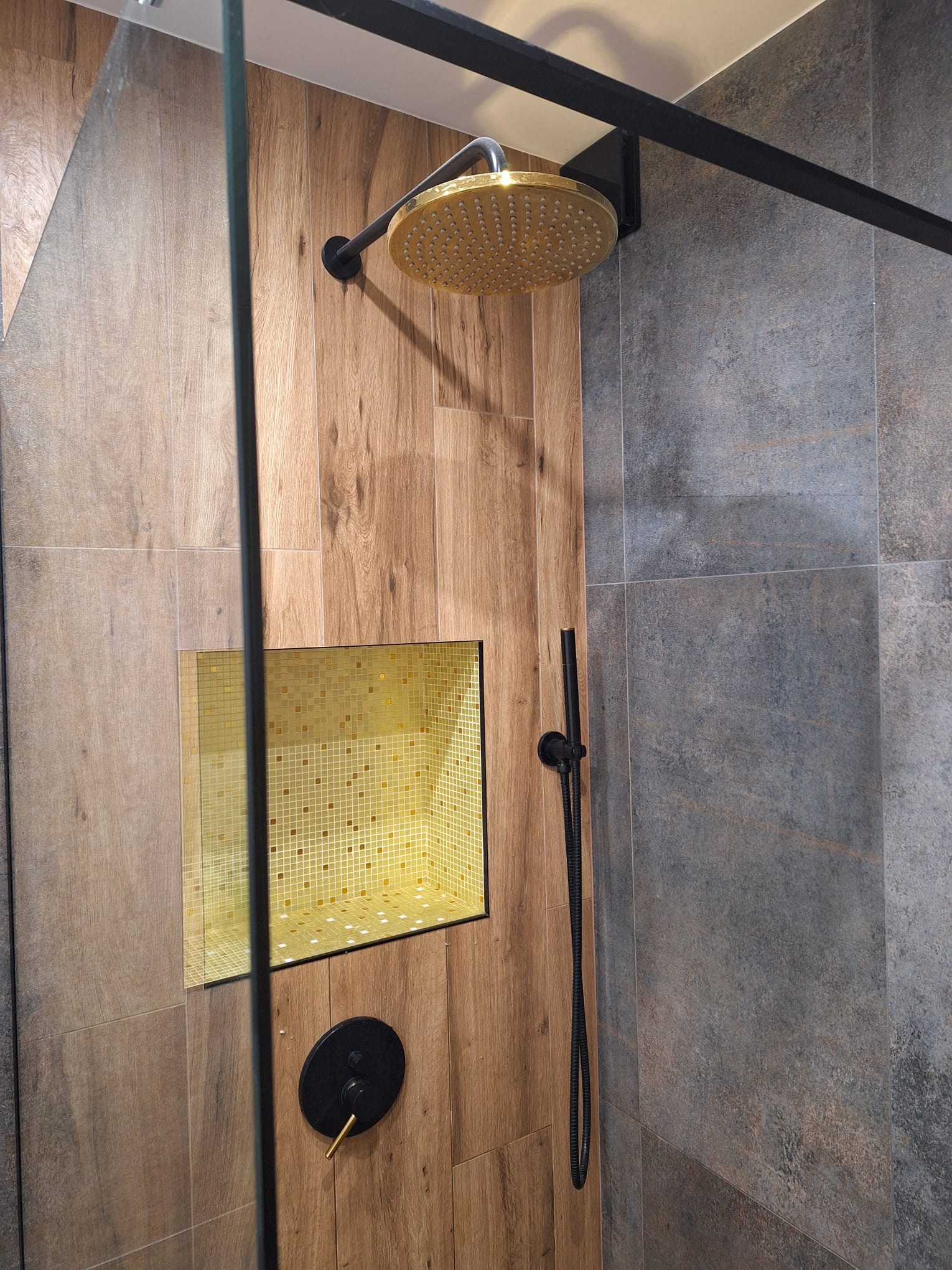 Nowoczesna kabina prysznicowa z drewnopodobnymi płytkami, złotą deszczownicą i wnęką wyłożoną mozaiką, widoczna czarna armatura.