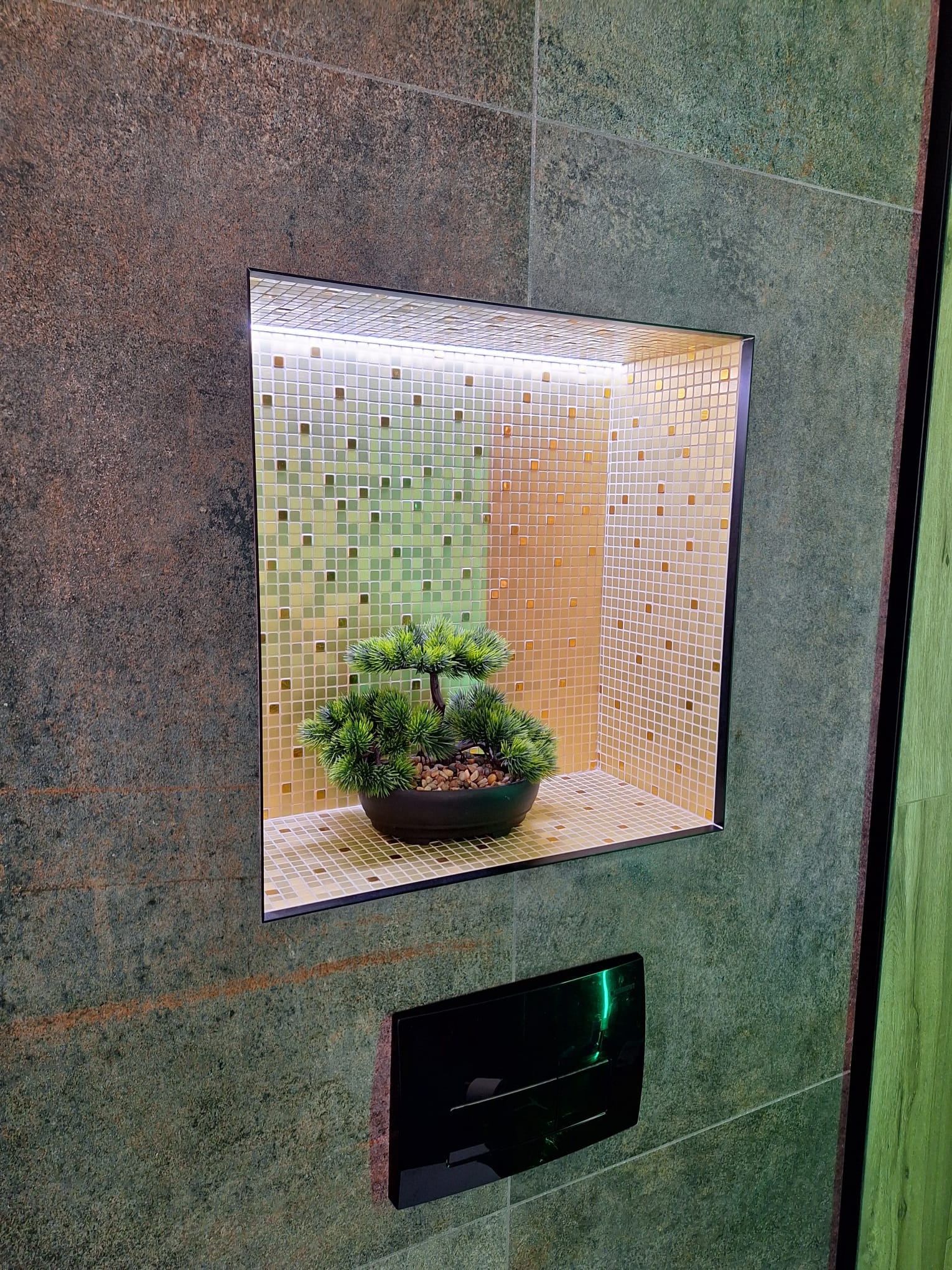 Wnęka w ścianie wyłożona mozaiką w odcieniach beżu i zieleni, podświetlona od góry, z dekoracyjnym bonsai w czarnej doniczce. Poniżej widoczny czarny panel sterowania.