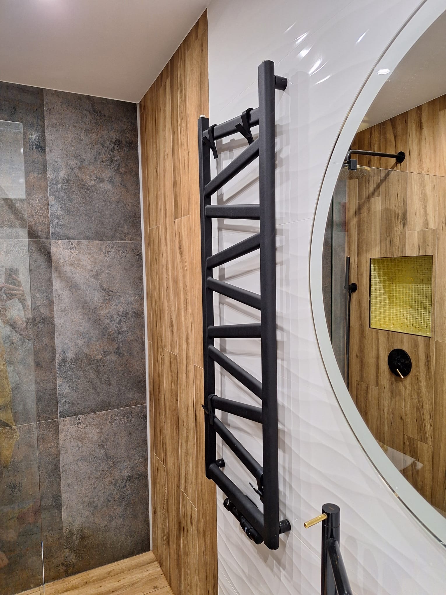 Nowoczesna łazienka z czarnym grzejnikiem drabinkowym, lustrem w owalnej ramie i okładzinami ściennymi imitującymi drewno i kamień.