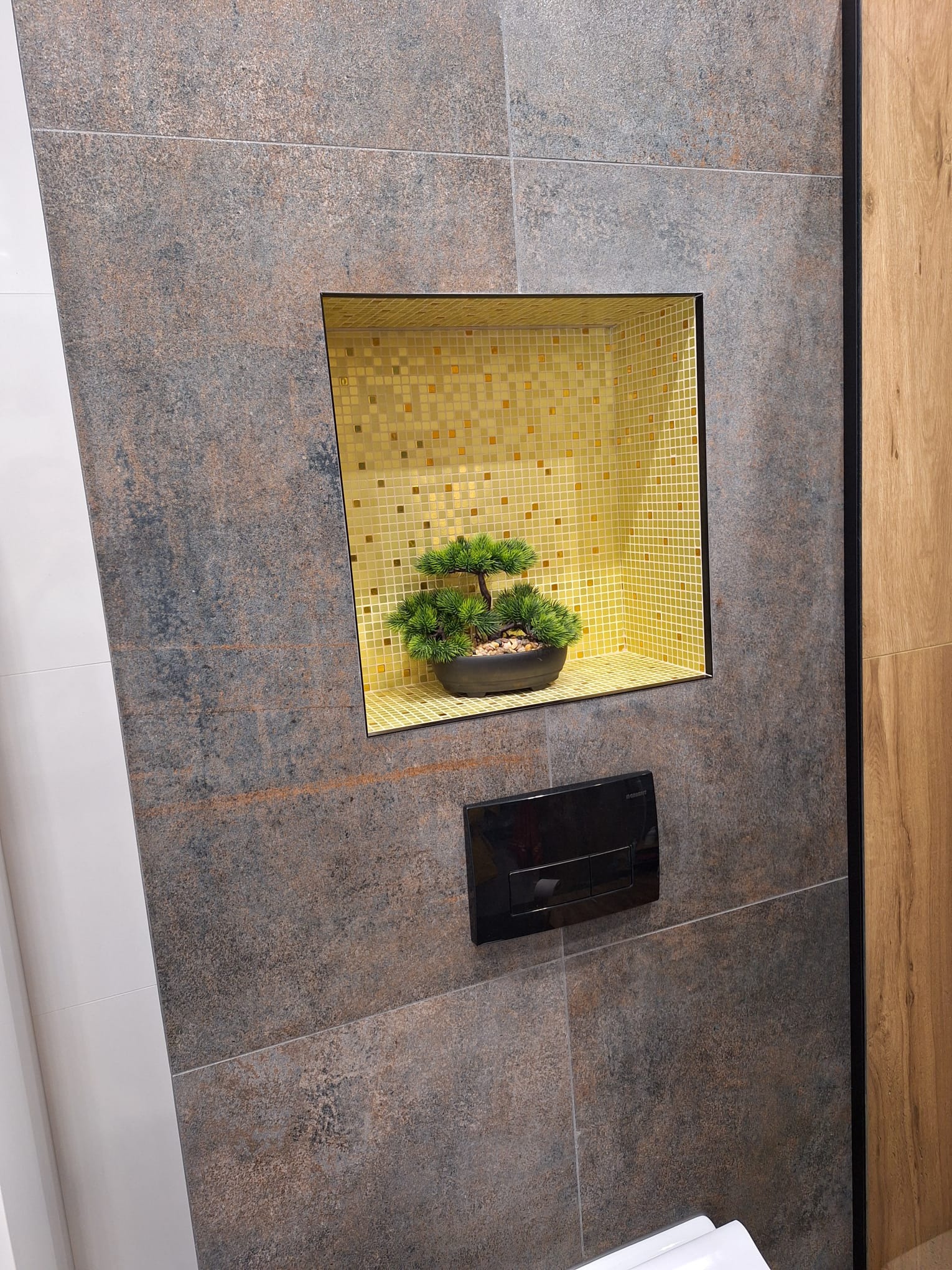 Wykończona łazienka z szarobrązowymi płytkami imitującymi kamień, wnęka z mozaiką w odcieniach złota i bonsai oraz czarny panel sterujący spłuczką WC.
