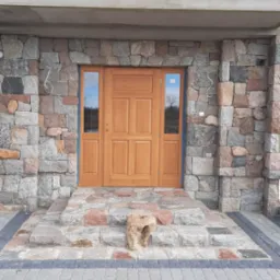 Wejście do domu z drewnianymi drzwiami, obłożone kamieniem naturalnym, z kamiennymi schodami i brukowanym chodnikiem, z surowym wykończeniem nadproża.