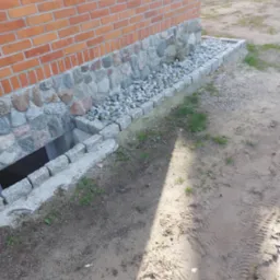 Fragment ściany budynku z cegły i kamienia z okienkiem piwnicznym, otoczony obramowaniem z kostki granitowej i wypełniony drobnymi kamieniami, widok z góry na piasek z odciśniętym śladem opon.