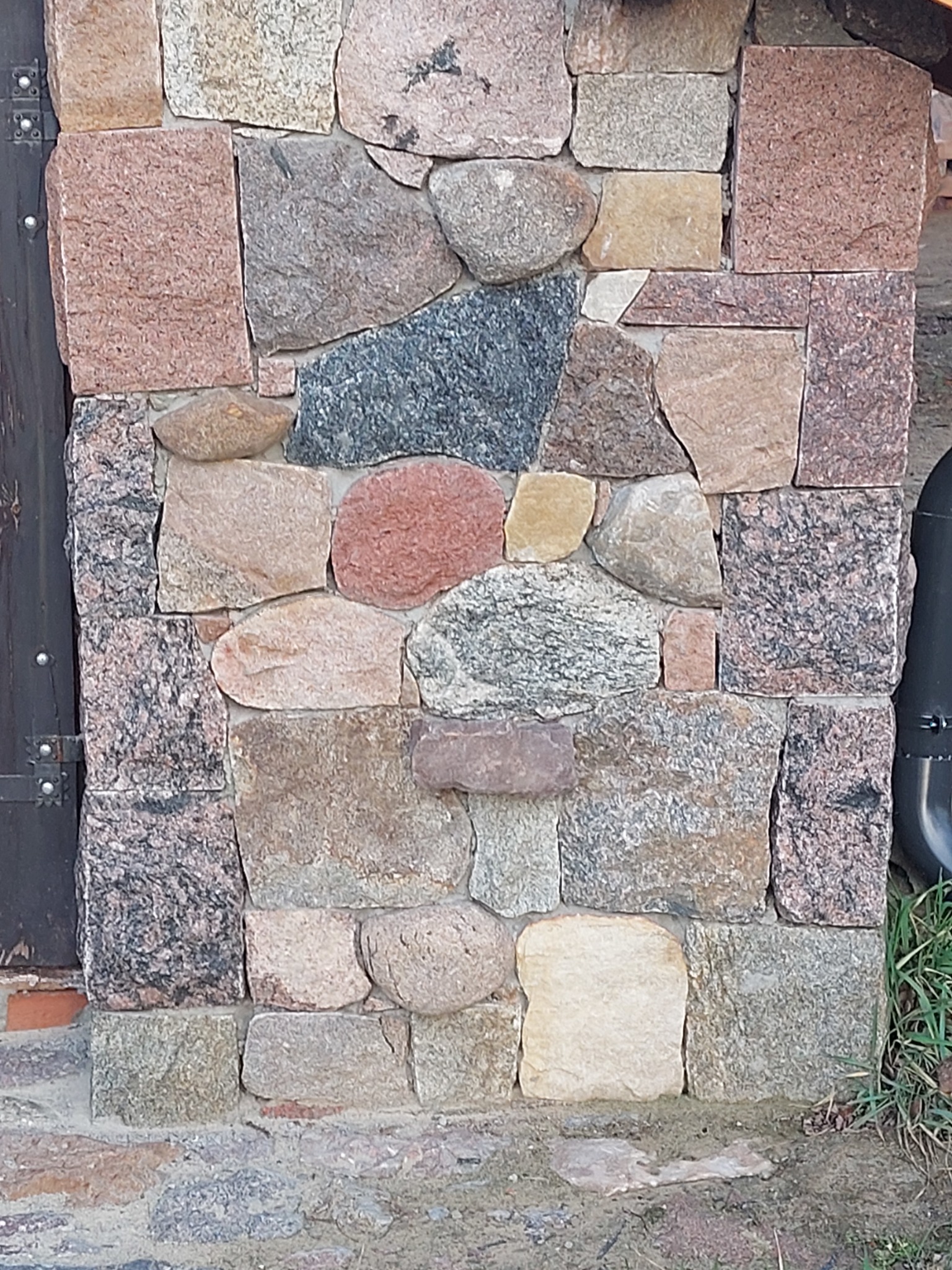 Fragment elewacji z nieregularnych kamieni o różnej fakturze i kolorystyce, z widocznymi elementami stolarki drzwiowej po lewej stronie i fragmentem instalacji po prawej.