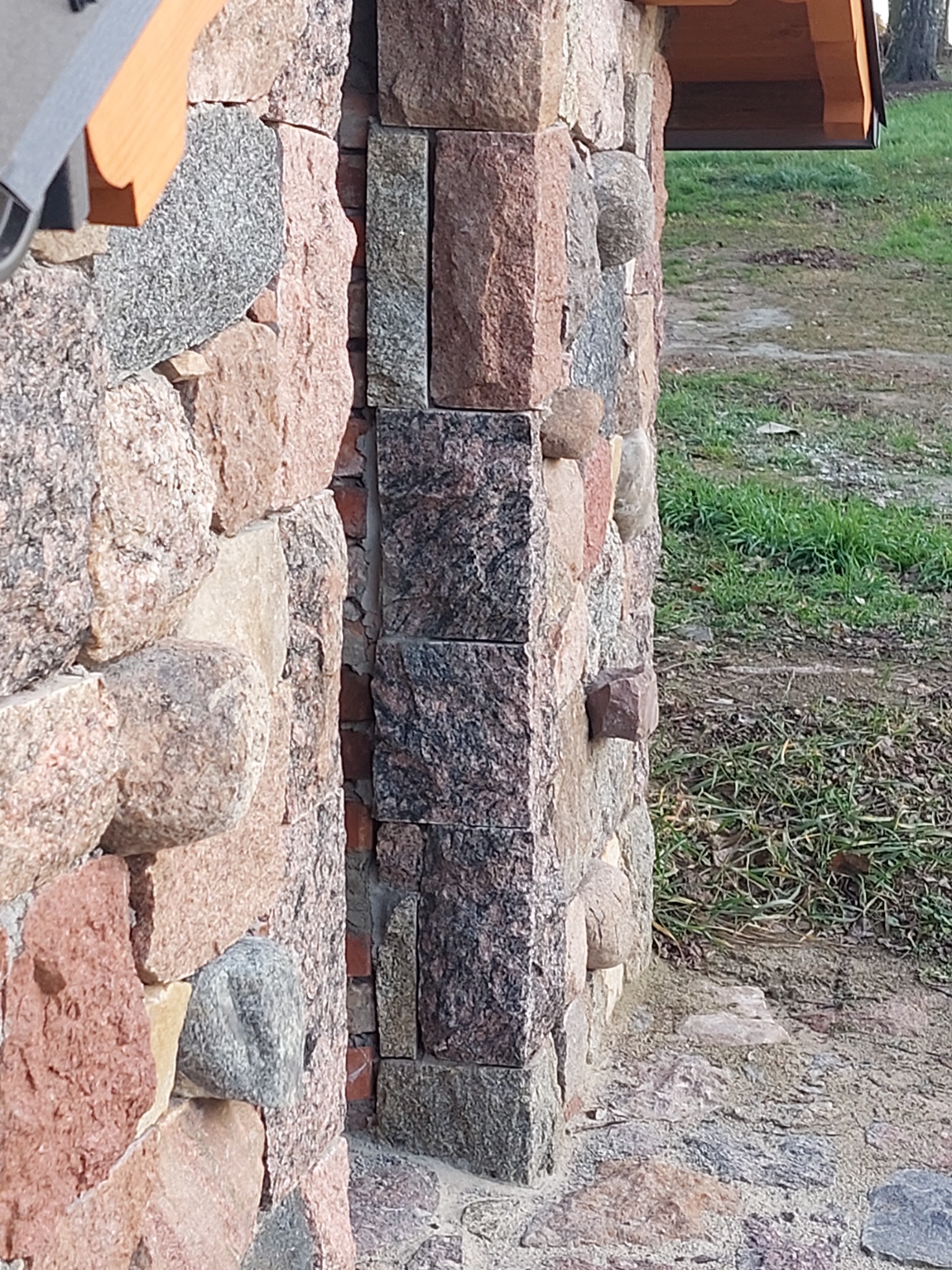 Fragment kamiennej ściany z narożnikiem wykończonym ciemniejszymi, regularnymi blokami, widok na trawnik w tle.