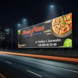 Billboard reklamowy pizzerii Street Pizza nocą, z rozmazanymi światłami samochodów na autostradzie. W tle budynki miejskie i latarnie uliczne.