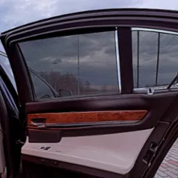 Wnętrze luksusowego samochodu z otwartymi drzwiami, widoczne rolety przeciwsłoneczne w oknach, drewniane wykończenie i jasna skórzana tapicerka.