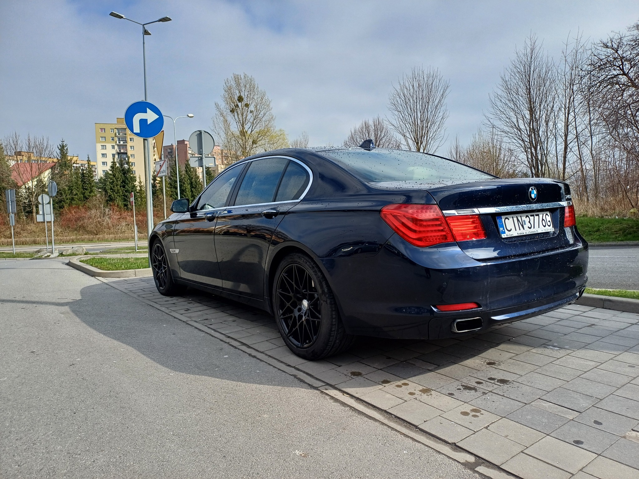 Granatowy samochód BMW serii 7 zaparkowany na chodniku z kostki brukowej, widok z tyłu, z widoczną tablicą rejestracyjną i czarnymi felgami.