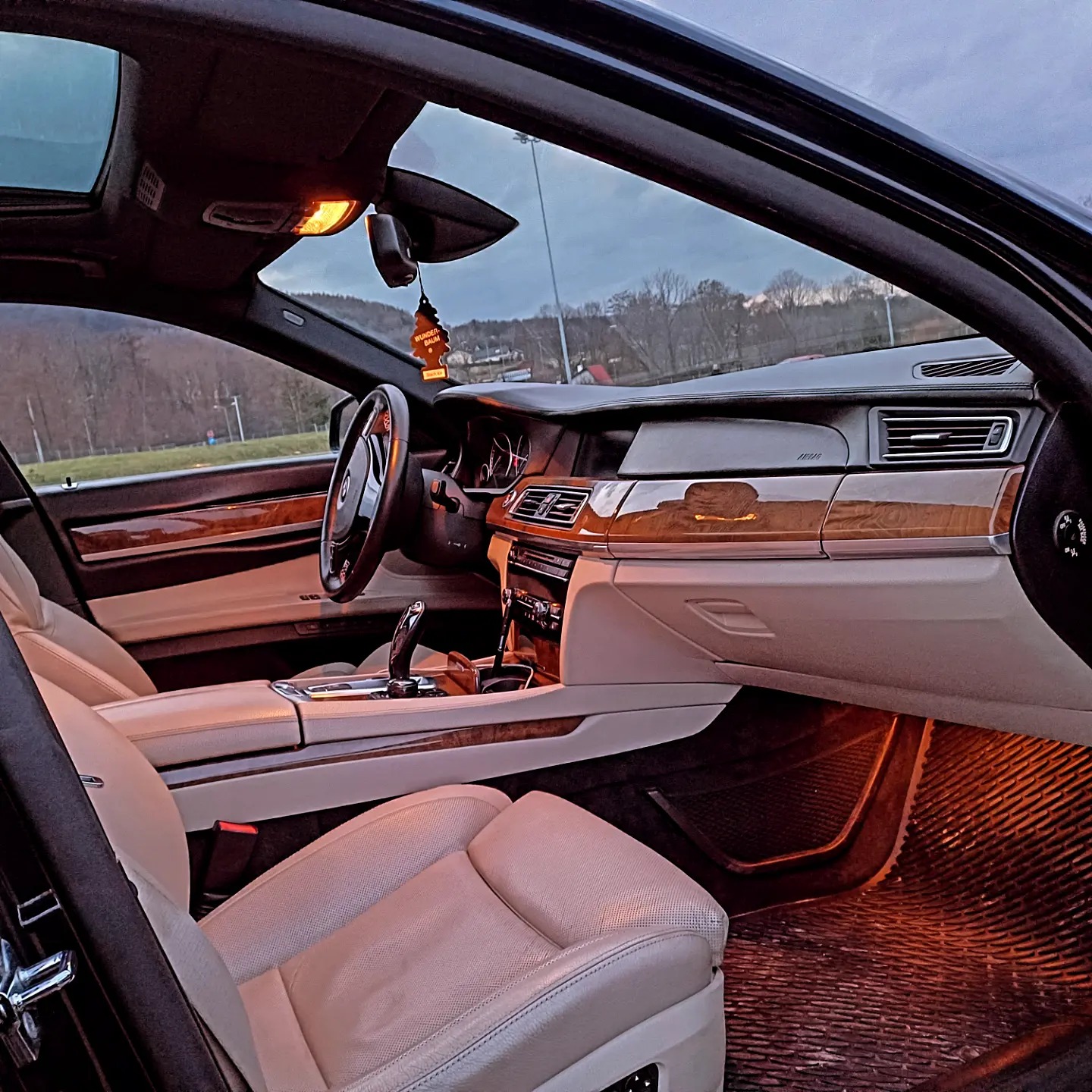 Wnętrze luksusowego samochodu z jasną skórzaną tapicerką i drewnianymi elementami wykończeniowymi, widok z zewnątrz przez otwarte drzwi, w tle krajobraz zalesiony.