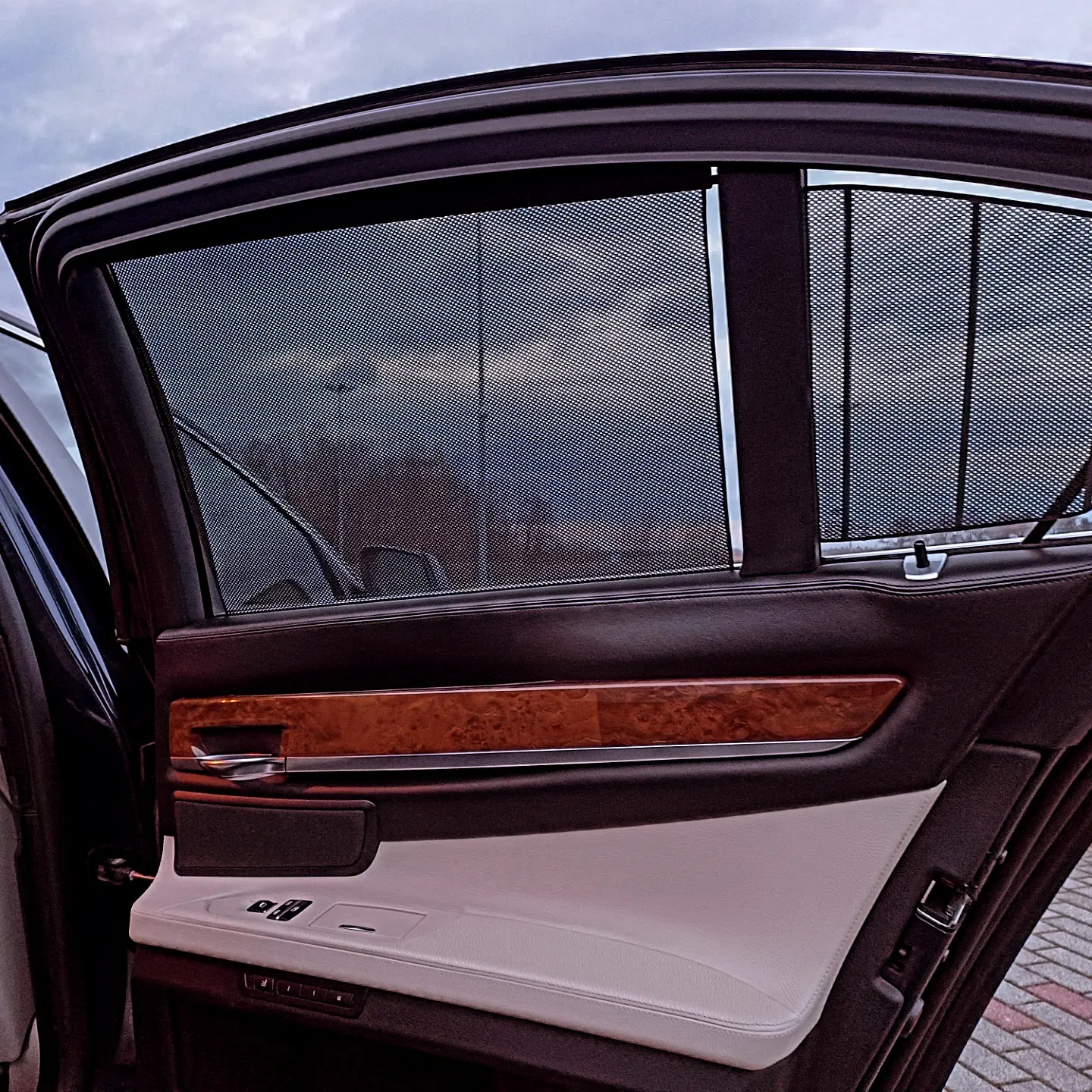 Wnętrze luksusowego samochodu z otwartymi drzwiami, widoczne rolety przeciwsłoneczne w oknach, drewniane wykończenie i jasna skórzana tapicerka.