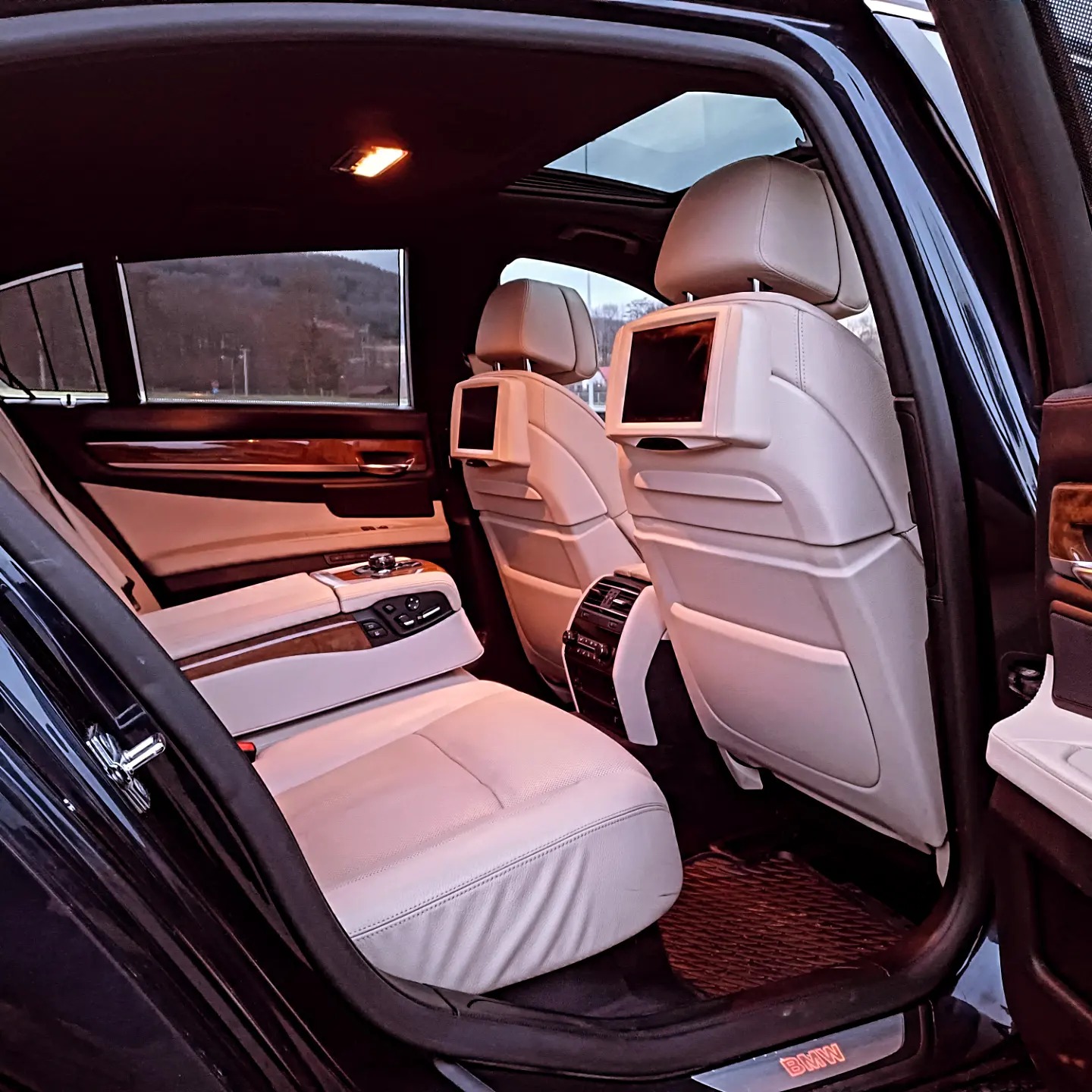 Luksusowe wnętrze samochodu z jasną skórzaną tapicerką, widoczne ekrany w zagłówkach tylnych siedzeń, podłokietnik z panelem sterowania i panoramiczny dach.