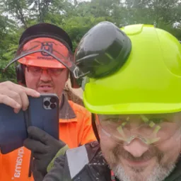 Dwóch pracowników w odzieży roboczej, hełmach i okularach ochronnych robi sobie selfie telefonem na tle zieleni.