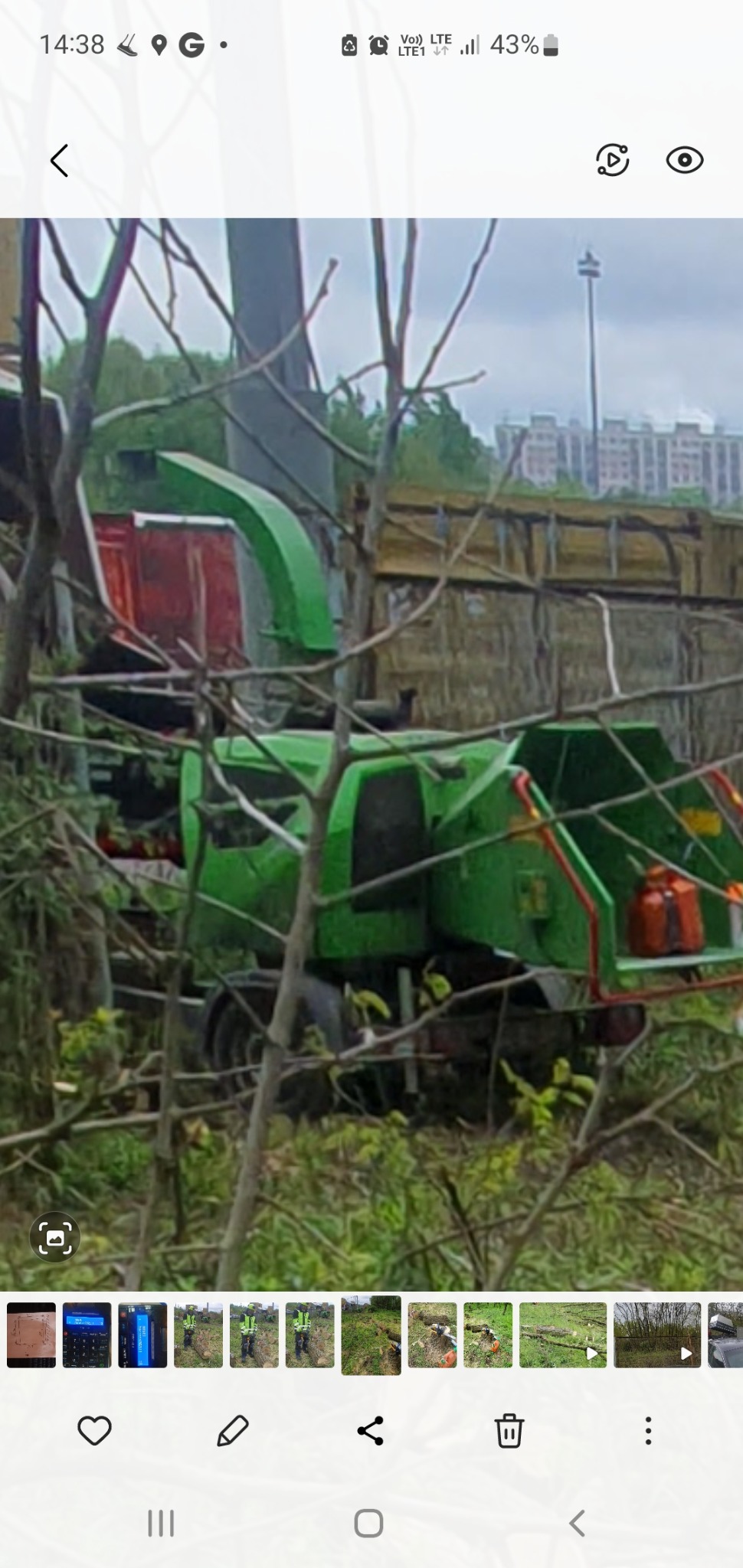 Zielona rębarka do gałęzi na tle drzew i budynku mieszkalnego, widoczna przez gałęzie.