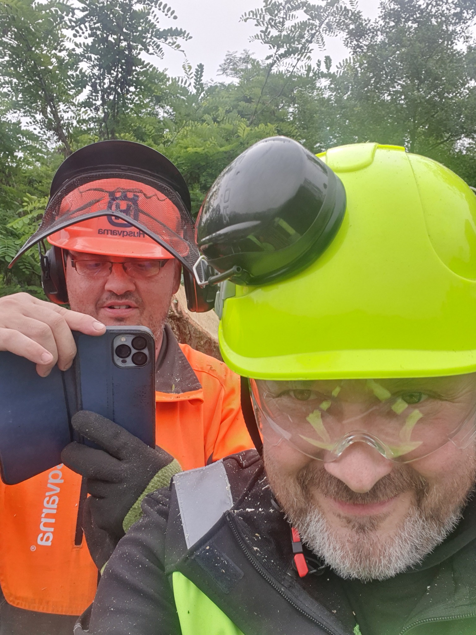 Dwóch pracowników w odzieży roboczej, hełmach i okularach ochronnych robi sobie selfie telefonem na tle zieleni.