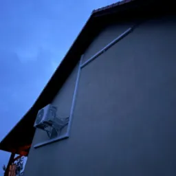 Zewnętrzna jednostka klimatyzacji zamontowana na ścianie budynku, z widocznymi przewodami w osłonie, sfotografowana o zmierzchu, z fragmentem dachu i nieba w tle.