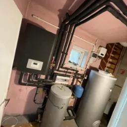 Piwnica z zainstalowanym systemem grzewczym: czarny piec, dwa srebrne zbiorniki na wodę, niebieski zbiornik wyrównawczy, rury w izolacji termicznej, okno w tle.