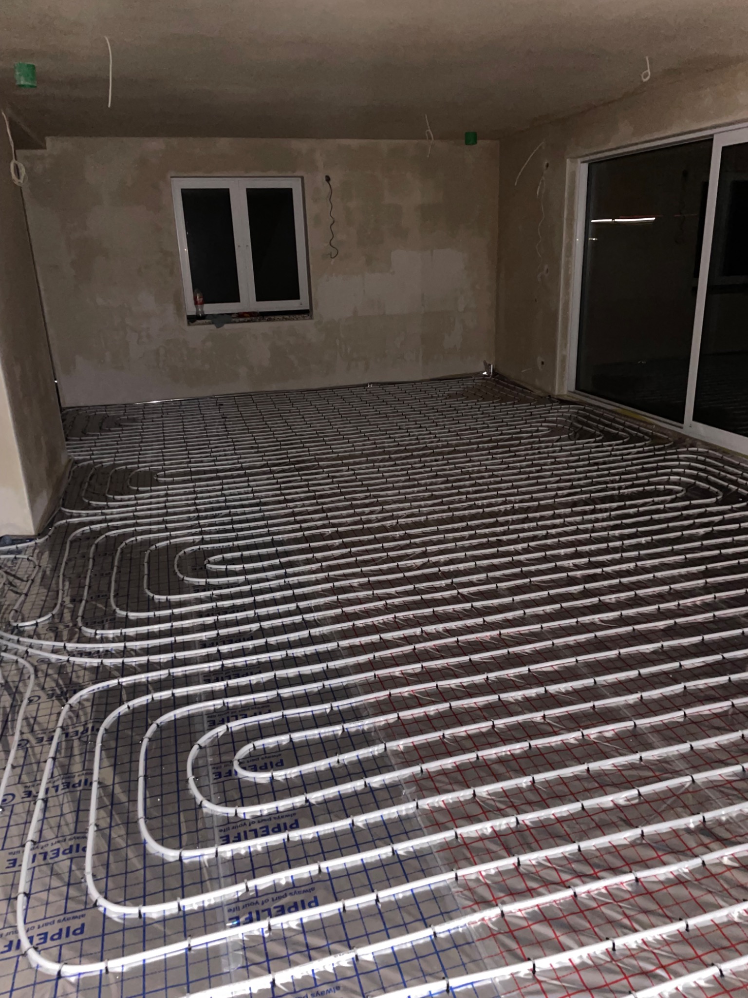 Rozłożone rury ogrzewania podłogowego na folii z nadrukiem PIPELIFE, widoczne okno i surowe ściany w trakcie remontu.