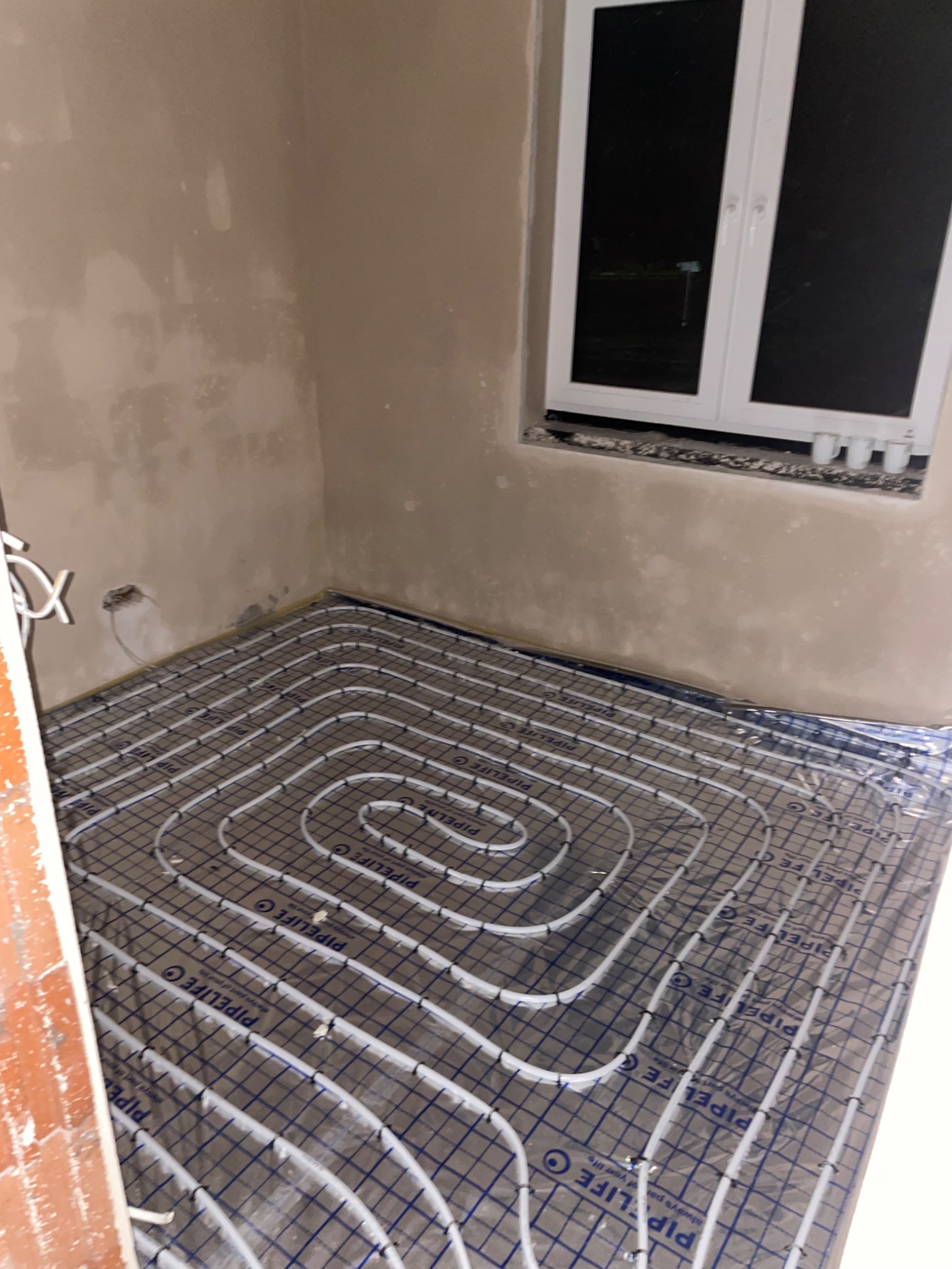 Białe rury systemu ogrzewania podłogowego ułożone spiralnie na folii z nadrukiem PIPELIFE w surowym wnętrzu z oknem.