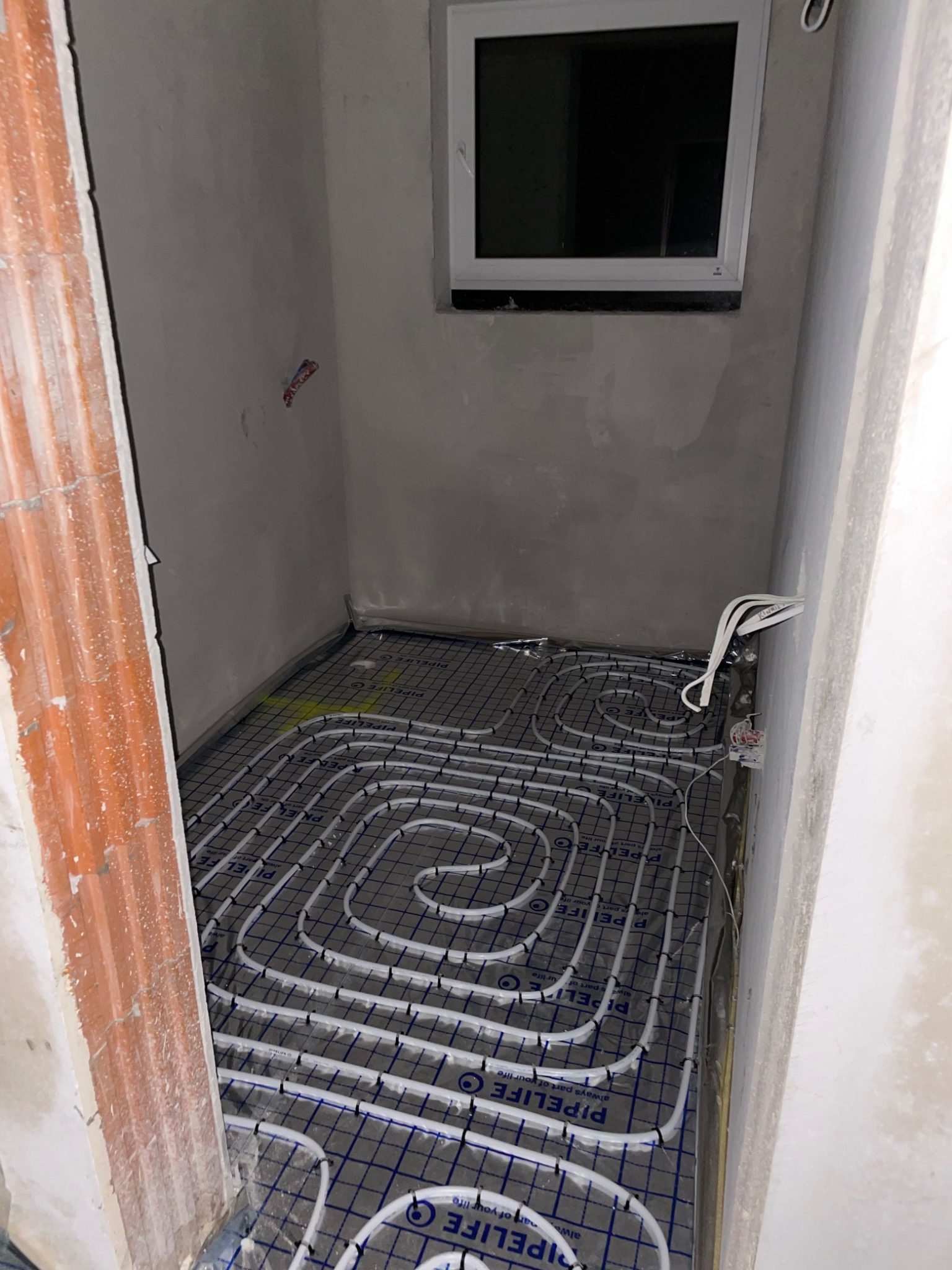 Rozłożona instalacja ogrzewania podłogowego z rurami Pipelife na siatce w surowym pomieszczeniu z oknem, widoczne przewody elektryczne i fragment ceglanej ściany.