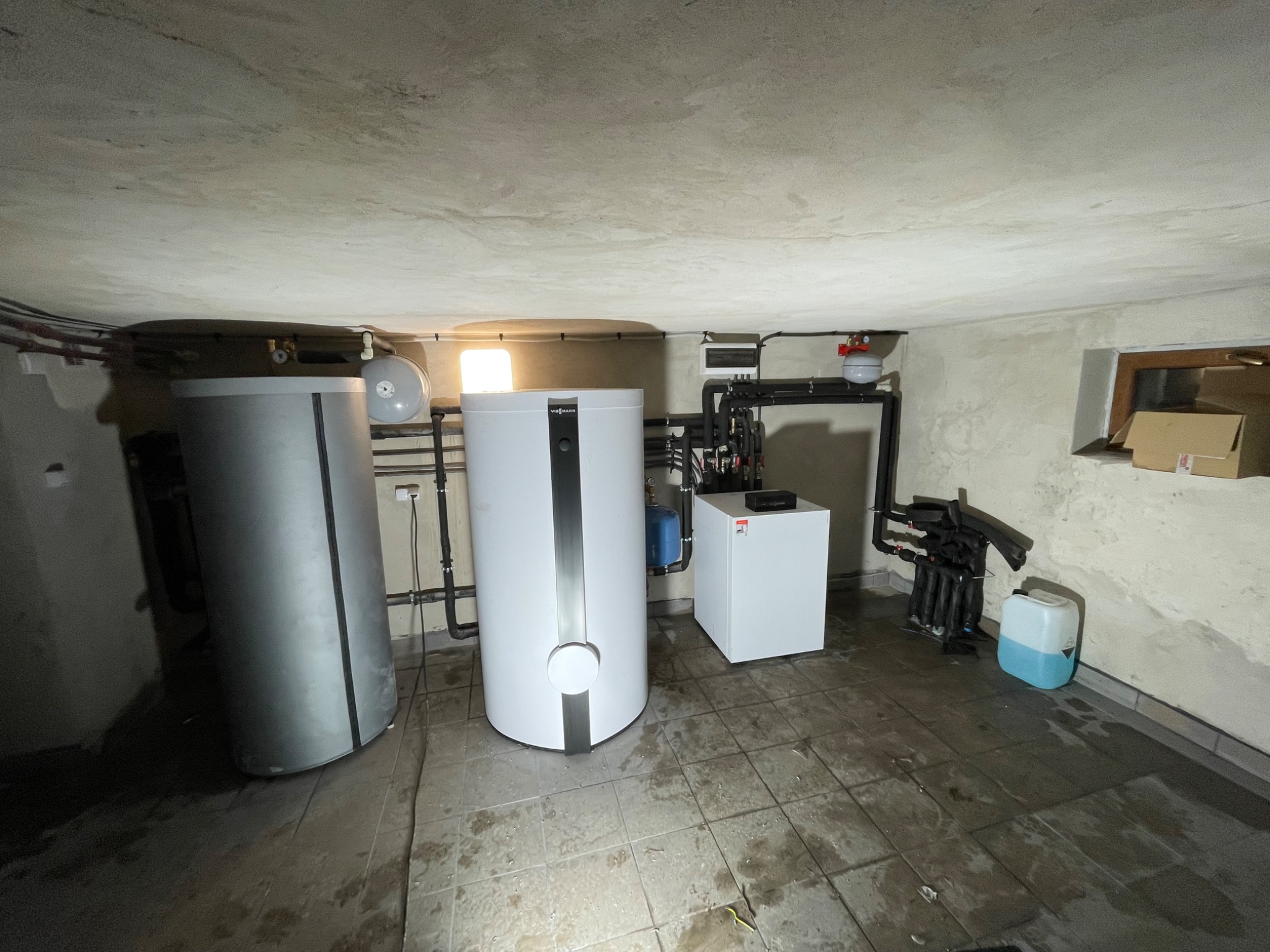 Piwnica z zainstalowanymi urządzeniami grzewczymi: srebrny zbiornik buforowy, biały zasobnik ciepłej wody z czarnym pionowym pasem oraz biały piec, połączone czarnymi rurami. Na podłodze płytki...