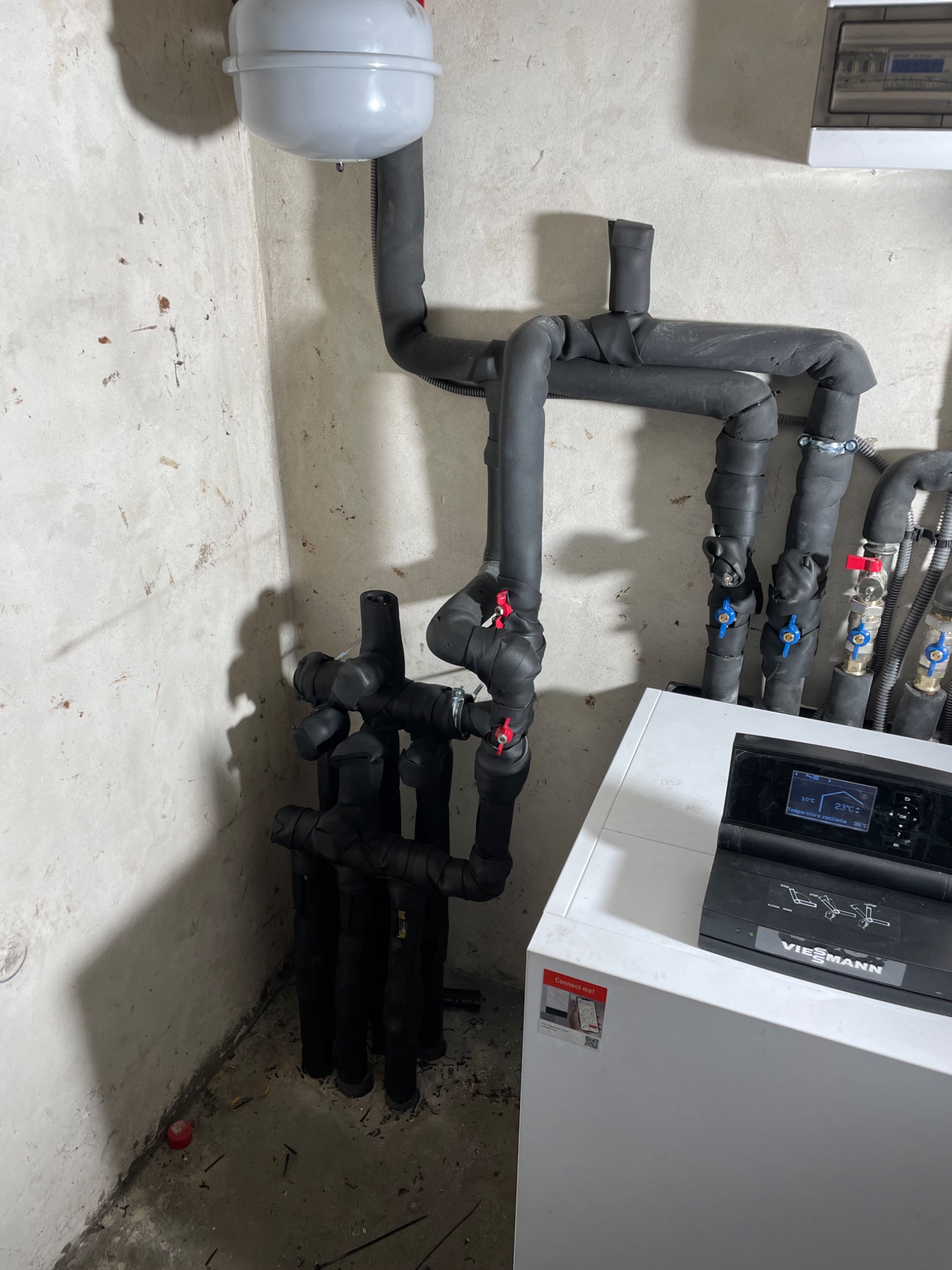 Instalacja grzewcza z rurami w izolacji, zaworami z czerwonymi i niebieskimi pokrętłami, zbiornikiem wyrównawczym i sterownikiem Viessmann na tle surowej ściany.