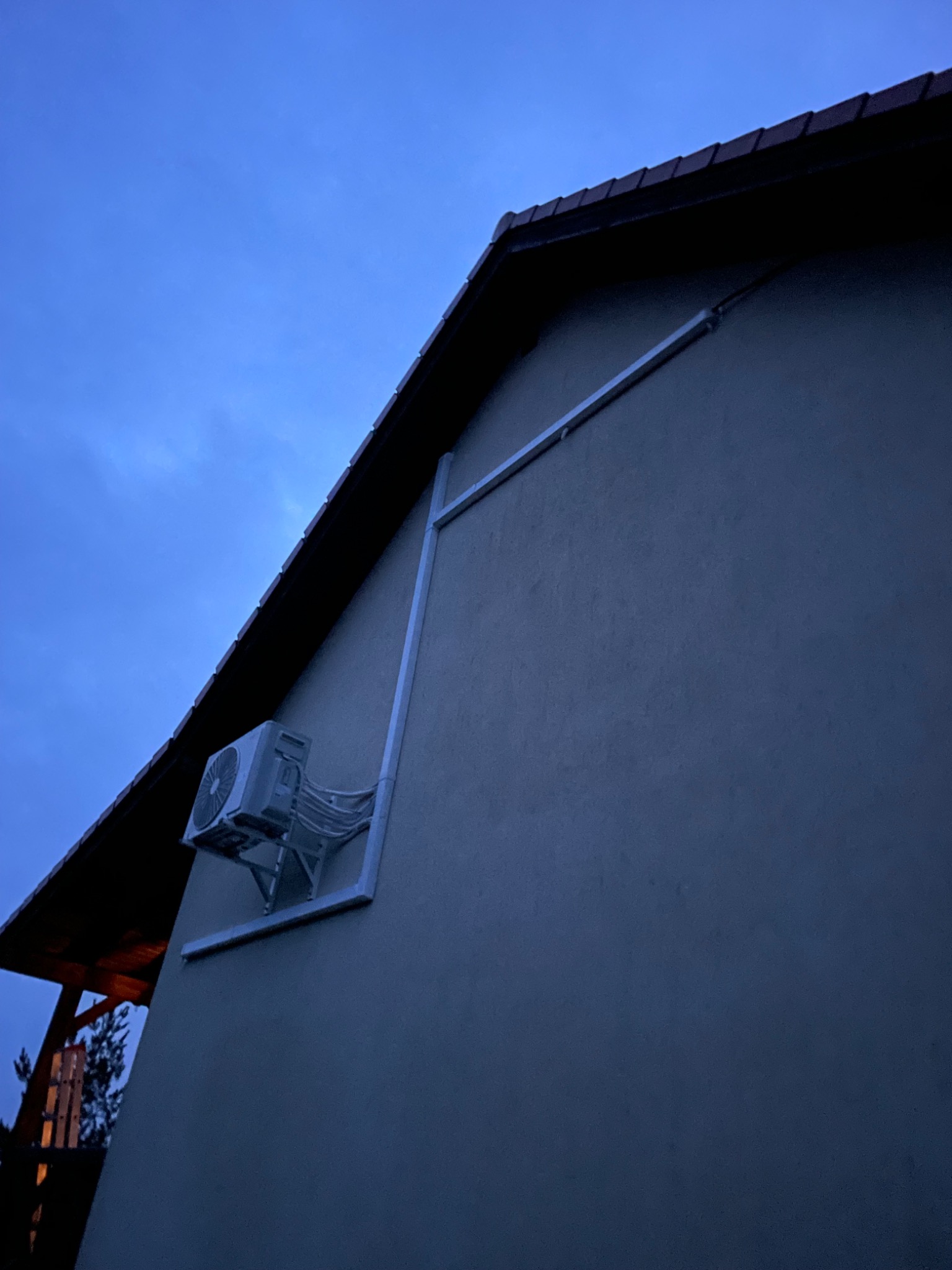 Zewnętrzna jednostka klimatyzacji zamontowana na ścianie budynku, z widocznymi przewodami w osłonie, sfotografowana o zmierzchu, z fragmentem dachu i nieba w tle.
