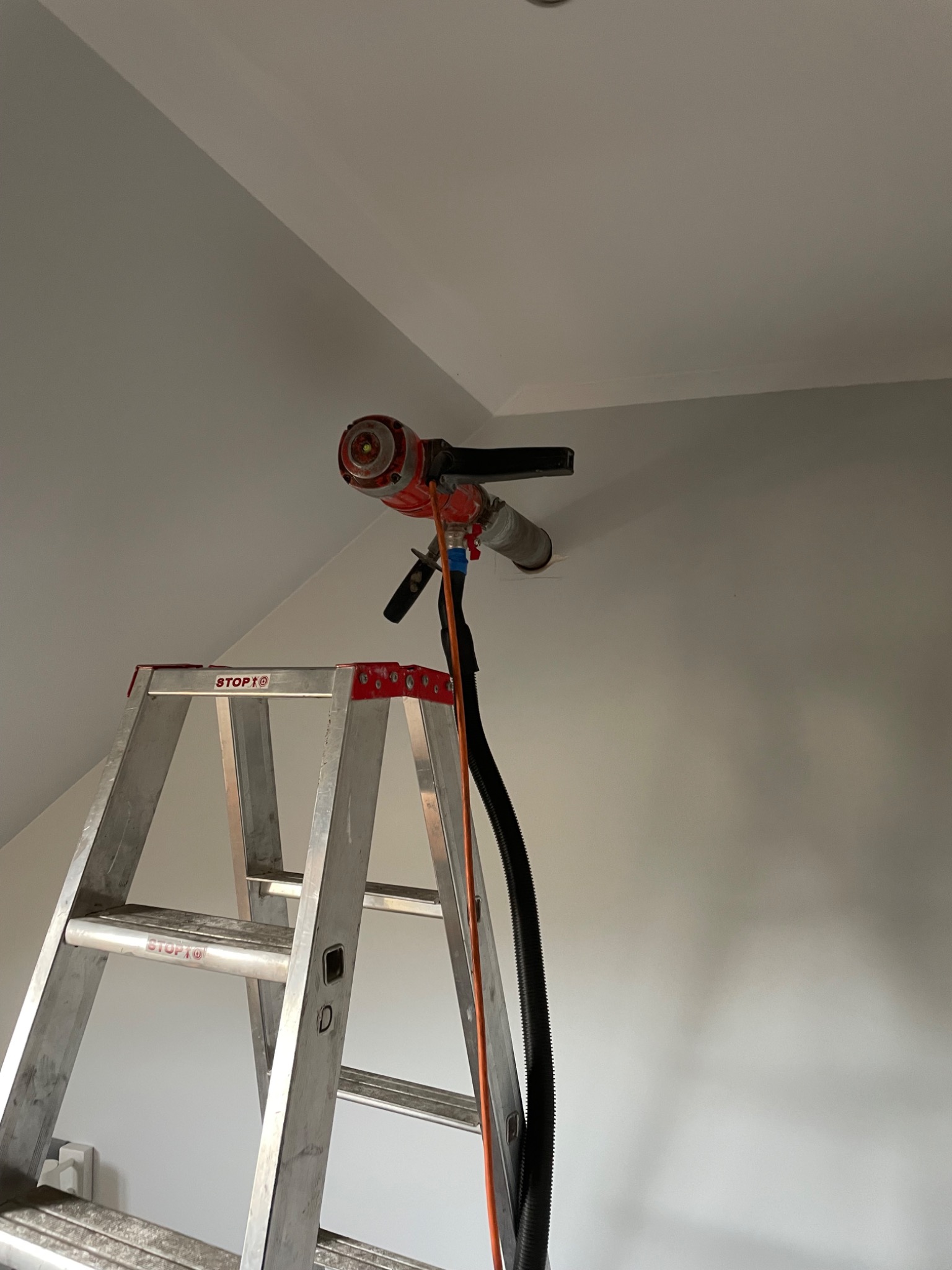 Wiertnica rdzeniowa zamontowana na ścianie nad aluminiową drabiną podczas wykonywania otworu wentylacyjnego w szarej ścianie pod skosem.