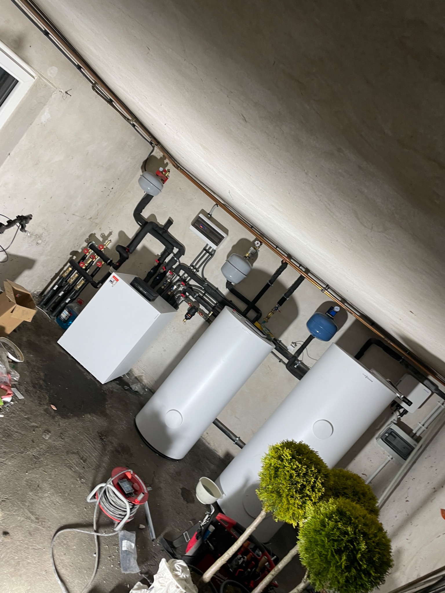 Kompletna instalacja grzewcza w kotłowni z dwoma białymi zbiornikami, kotłem, orurowaniem, osprzętem, oraz dekoracyjnymi roślinami w doniczkach na podłodze.