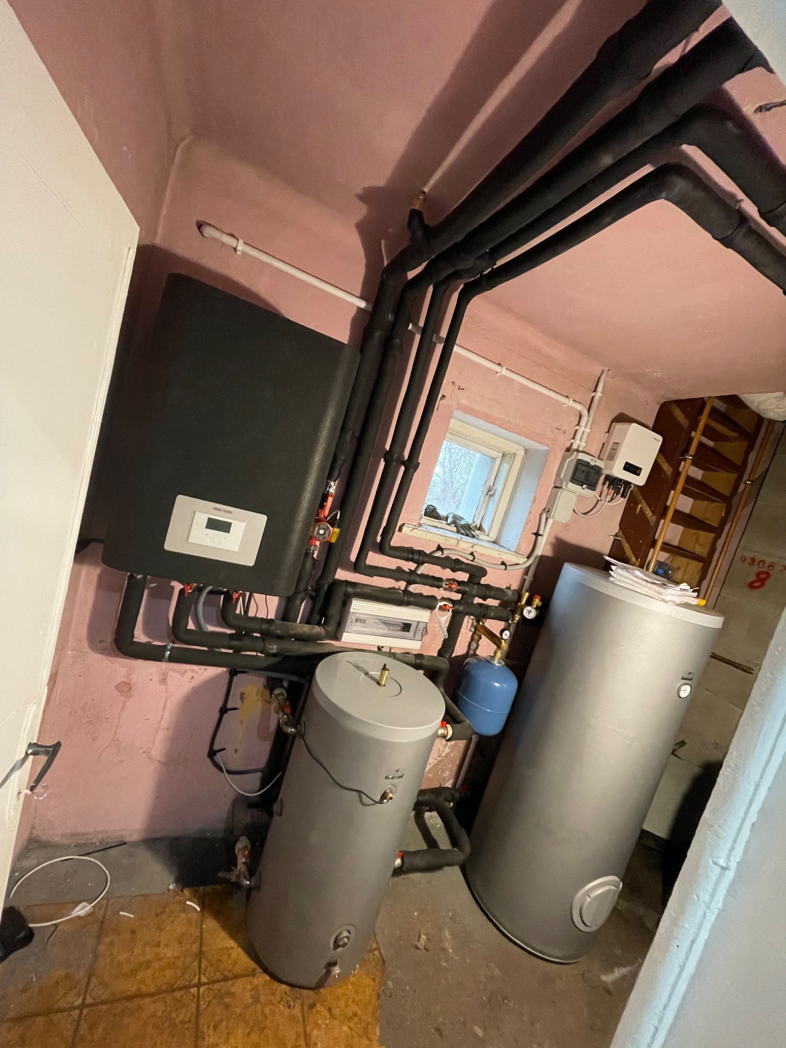 Piwnica z zainstalowanym systemem grzewczym: czarny piec, dwa srebrne zbiorniki na wodę, niebieski zbiornik wyrównawczy, rury w izolacji termicznej, okno w tle.