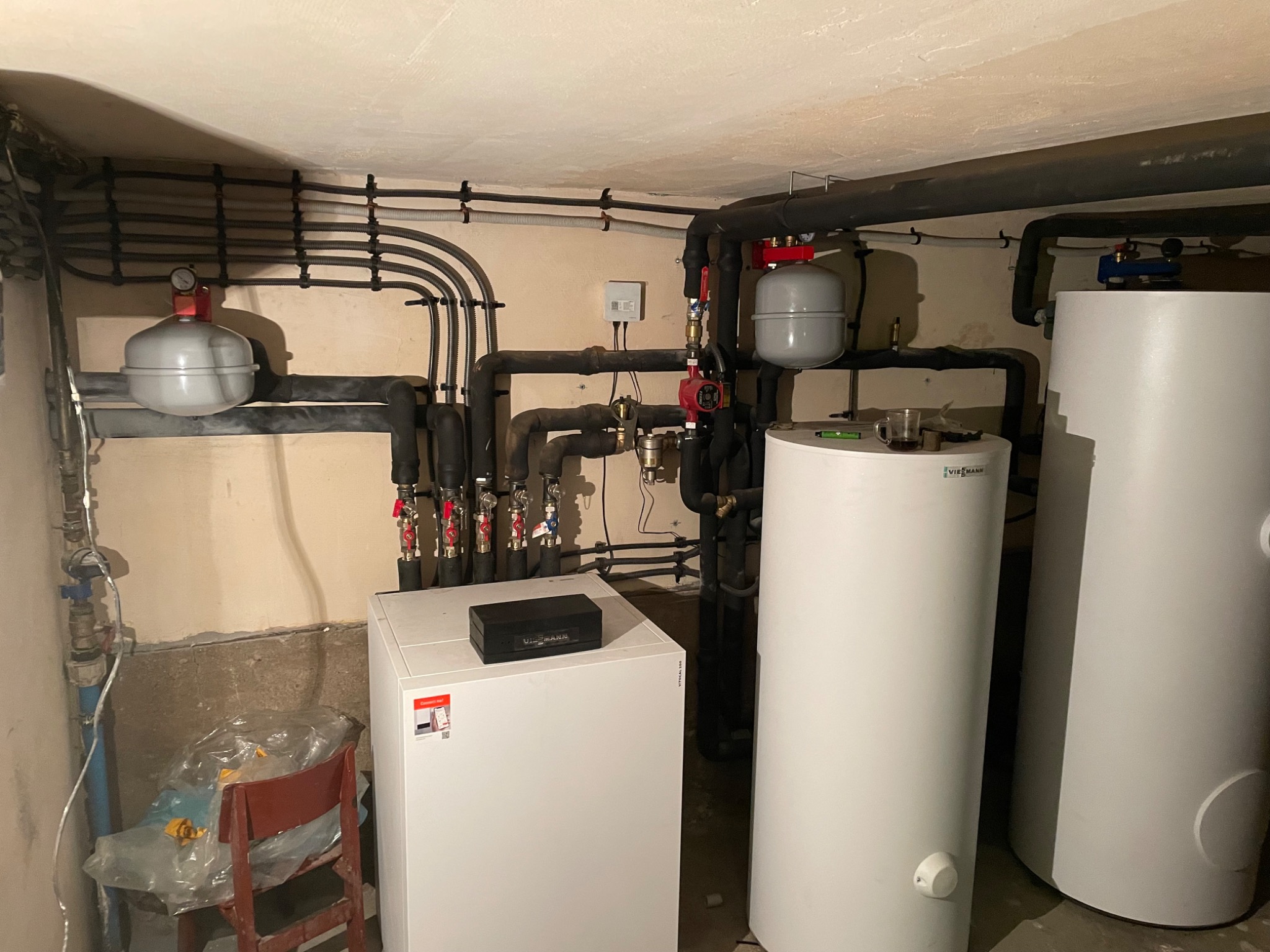 Instalacja grzewcza w piwnicy z rurami, zbiornikami wyrównawczymi i kotłem Viessmann. Na zbiorniku ciepłej wody stoi niedopita herbata w szklance, a obok kotła stoi krzesło z folią ochronną.