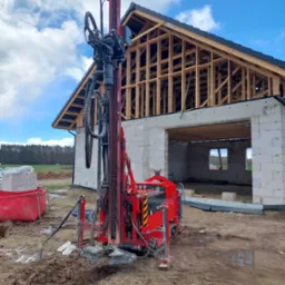 Czerwona wiertnica na gąsienicach ustawiona na placu budowy domu w stanie surowym otwartym, widoczne przewody i elementy robocze, w tle las i błękitne niebo z chmurami.