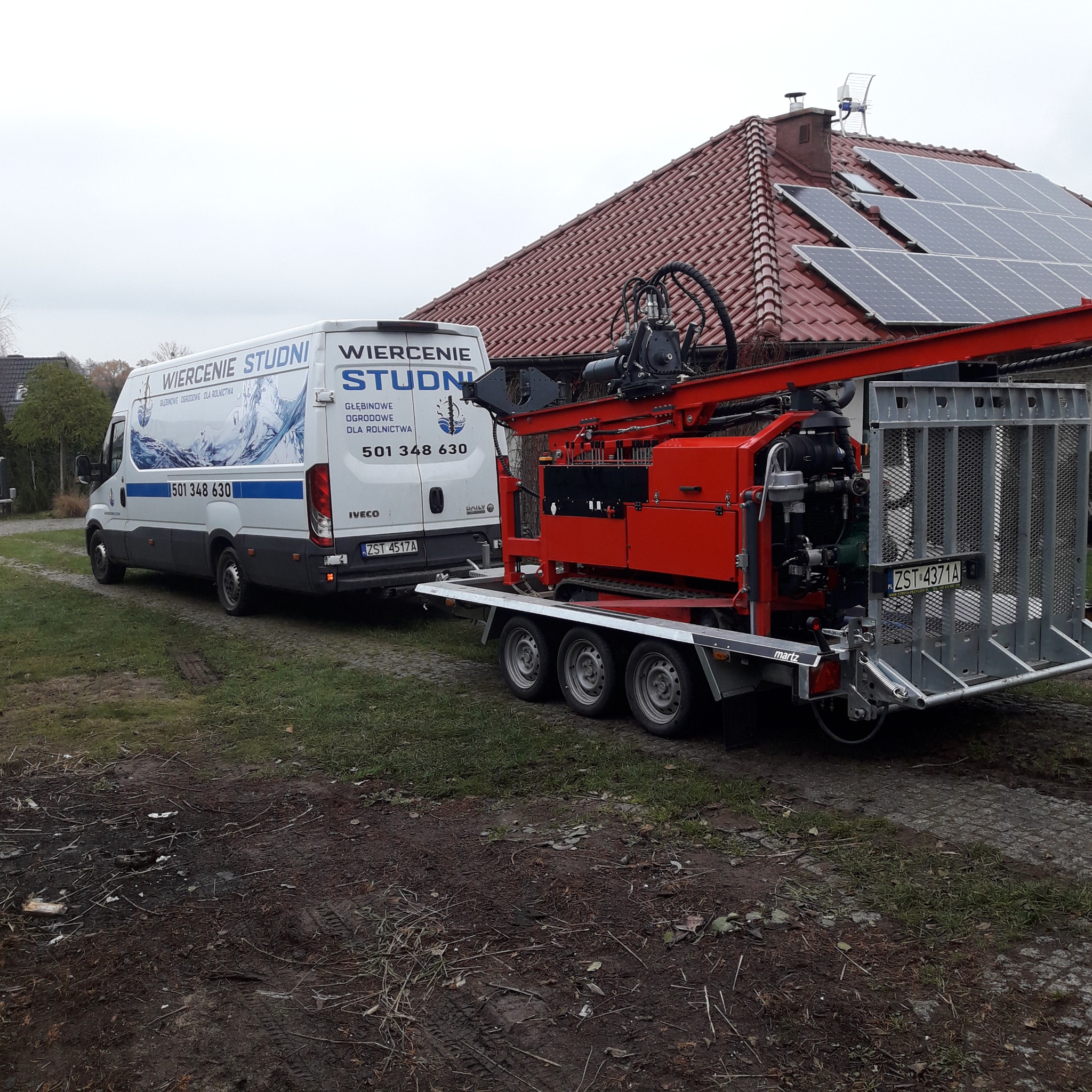 Czerwona wiertnica do studni głębinowych na przyczepie, zaparkowana obok białego samochodu dostawczego z reklamą wiercenia studni, na tle domu z panelami słonecznymi.