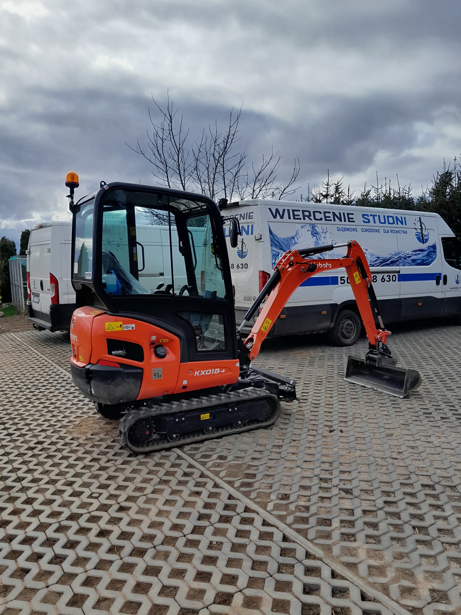 Pomarańczowa mini koparka Kubota KX018-4 na betonowej nawierzchni ażurowej, z furgonetką z napisem 'Wiercenie Studni' w tle.