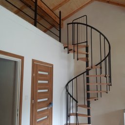 Usługi remontow Budowlane Leszek Wiśniewski - Wnętrze z drewnianym stropem, spiralnymi schodami z czarną balustradą i drewnianymi stopniami. Widoczne dwoje drzwi z drewnianymi ościeżnicami.
