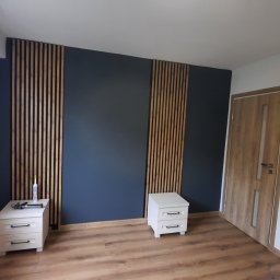 Usługi remontow Budowlane Leszek Wiśniewski - Nowoczesne wykończenie wnętrza sypialni: ciemna ściana z drewnianymi lamelami, panele podłogowe, białe szafki nocne i drzwi z drewnianym fornirem.