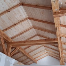 Usługi remontow Budowlane Leszek Wiśniewski - Drewniany szkielet dachu z boazerią, widok z dołu. Elementy konstrukcyjne z jasnego drewna, sufit z paneli. Fragment ściany z płyt gipsowo-kartonowych w lewym dolnym rogu.