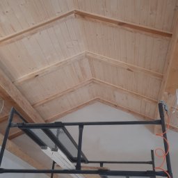 Usługi remontow Budowlane Leszek Wiśniewski - Drewniana boazeria na poddaszu, widok z rusztowania. Jasne drewno, skośny sufit. W tle biała ściana. Pomarańczowy przewód pneumatyczny na rusztowaniu.