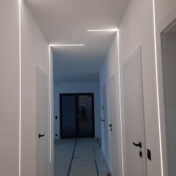 Usługi remontow Budowlane Leszek Wiśniewski - Biały korytarz z nowoczesnym oświetleniem LED wzdłuż ścian i sufitu. Podłoga zabezpieczona kartonem. Widoczne trzy białe drzwi z czarnymi klamkami.