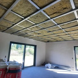 Usługi remontow Budowlane Leszek Wiśniewski - Poddasze z izolacją z wełny mineralnej w metalowym stelażu, widoczne rolki materiału Rockwool. Duże okna w tle, surowy beton na podłodze. Ocieplenie w trakcie prac.
