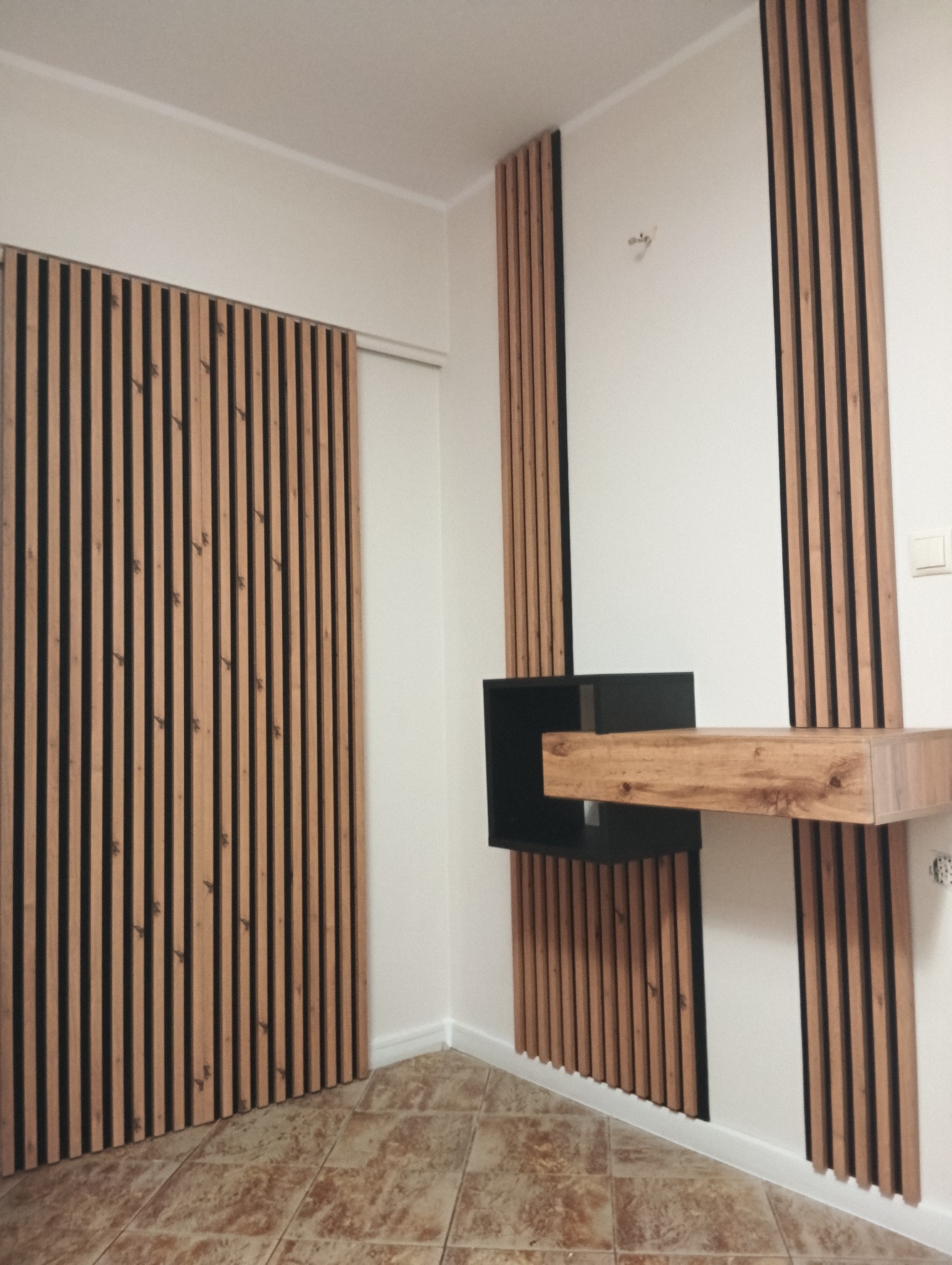 Nowoczesne wykończenie wnętrza z drewnianymi lamelami na ścianie i drzwiach, uzupełnione czarną szafką i drewnianą półką. Brązowa podłoga z płytek.