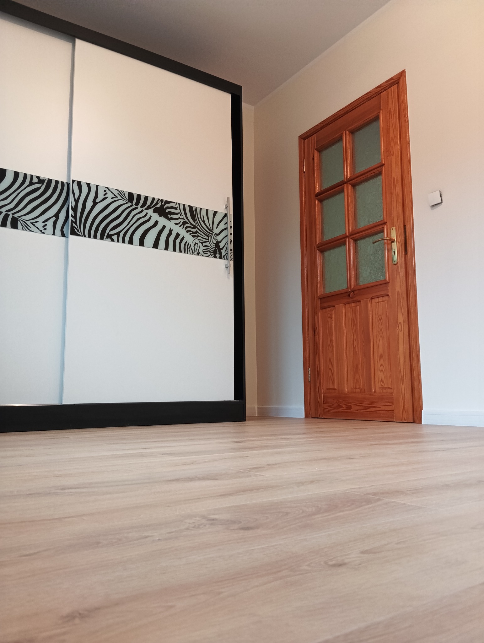 Jasne panele podłogowe, drewniane drzwi z szybkami i szafa przesuwna z motywem zebry w nowocześnie wykończonym wnętrzu.