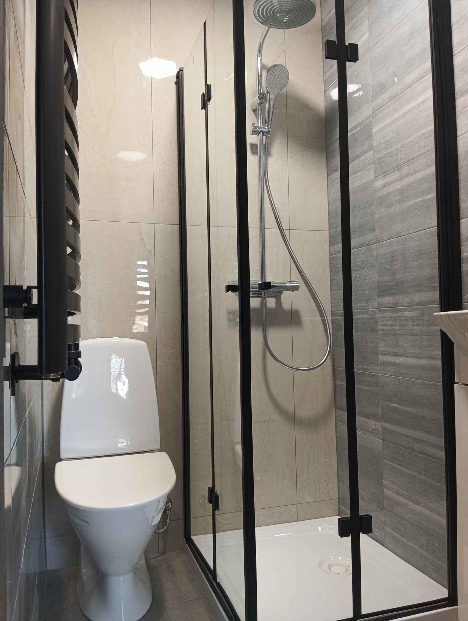 Wykończona łazienka z kabiną prysznicową typu walk-in z czarnymi profilami, toaletą i grzejnikiem drabinkowym. Płytki w odcieniach szarości i beżu.