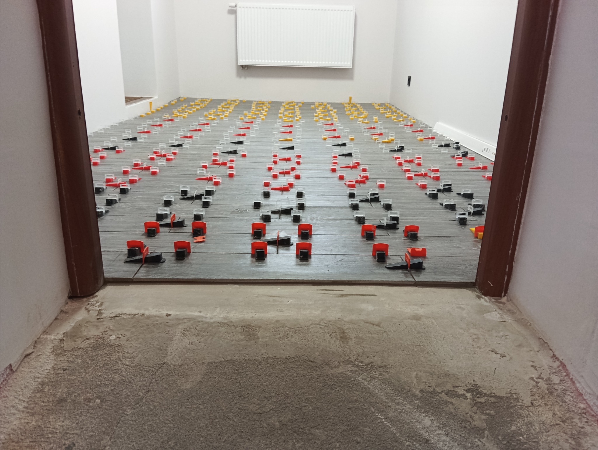 Układanie płytek podłogowych z użyciem systemu poziomowania; widok z progu, szare płytki, kolorowe kliny i klipsy, białe ściany, grzejnik na ścianie w tle.