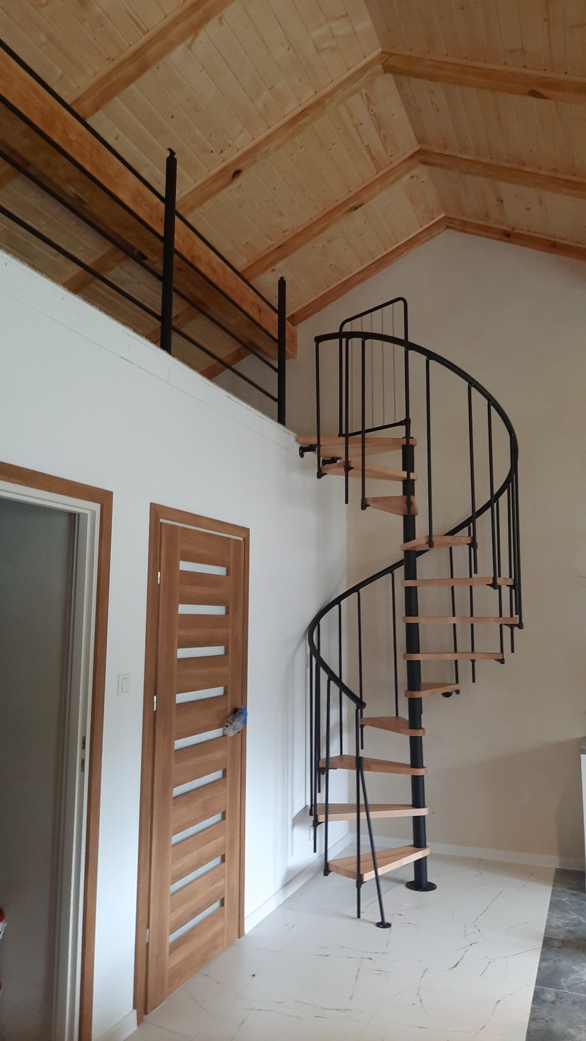 Wnętrze z drewnianym stropem, spiralnymi schodami z czarną balustradą i drewnianymi stopniami. Widoczne dwoje drzwi z drewnianymi ościeżnicami.