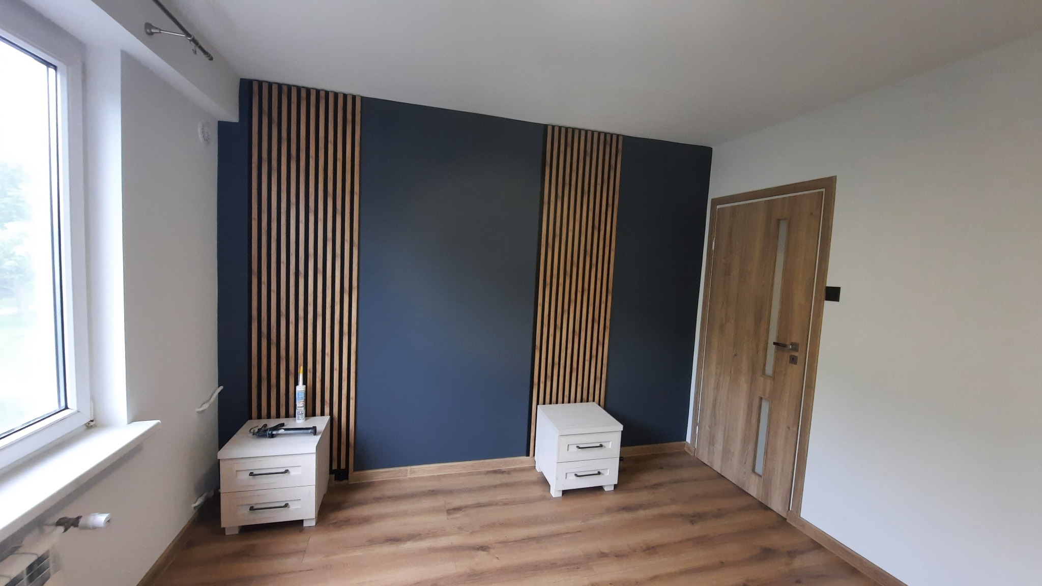 Nowoczesne wykończenie wnętrza sypialni: ciemna ściana z drewnianymi lamelami, panele podłogowe, białe szafki nocne i drzwi z drewnianym fornirem.