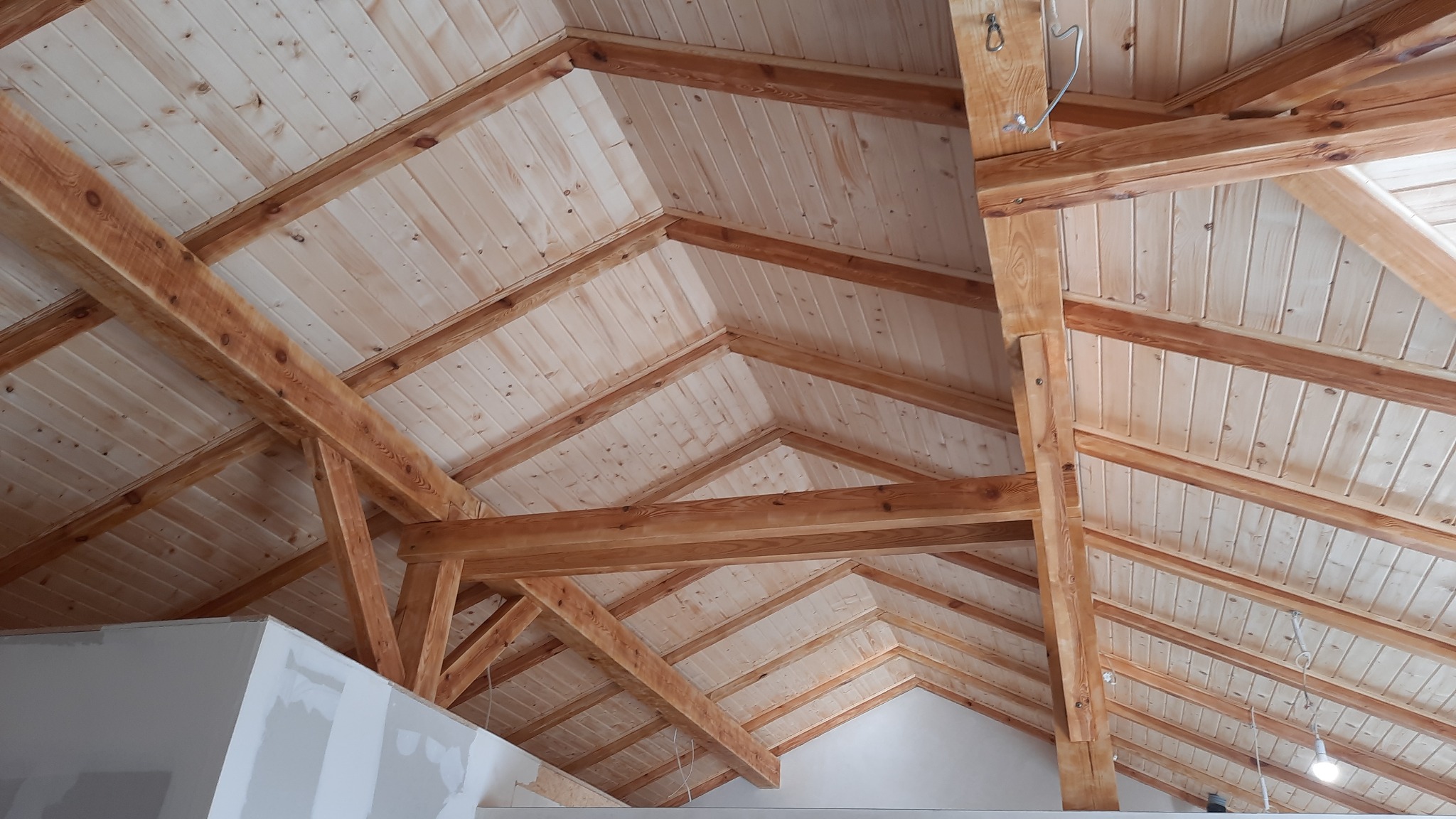 Drewniany szkielet dachu z boazerią, widok z dołu. Elementy konstrukcyjne z jasnego drewna, sufit z paneli. Fragment ściany z płyt gipsowo-kartonowych w lewym dolnym rogu.