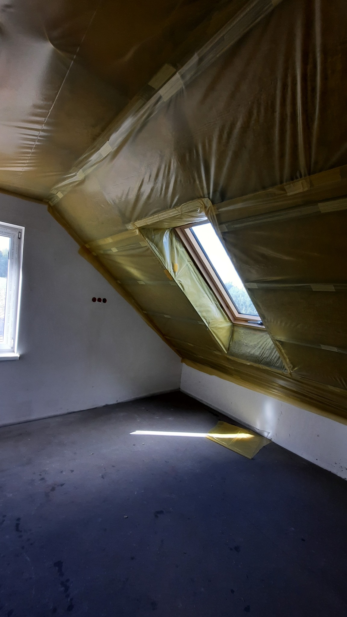 Poddasze z oknem dachowym w trakcie ocieplania, folia paroizolacyjna zabezpiecza skośny sufit. Widoczne trzy otwory na gniazdka elektryczne.