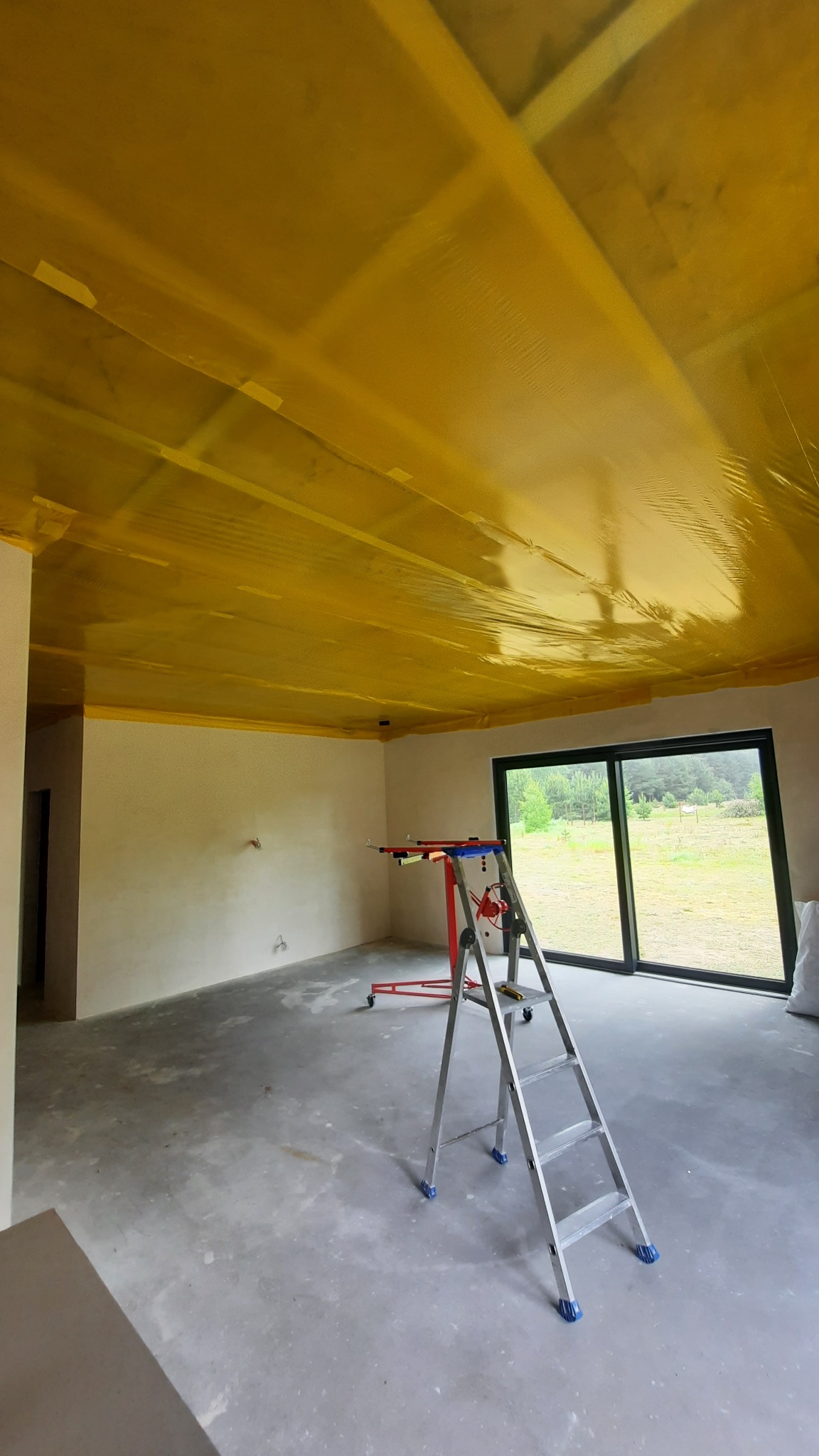 Wnętrze w trakcie prac przygotowawczych: sufit zabezpieczony folią, drabina i podnośnik do płyt. Widok na okno z zielenią w tle. Przygotowanie do dalszych prac wykończeniowych.