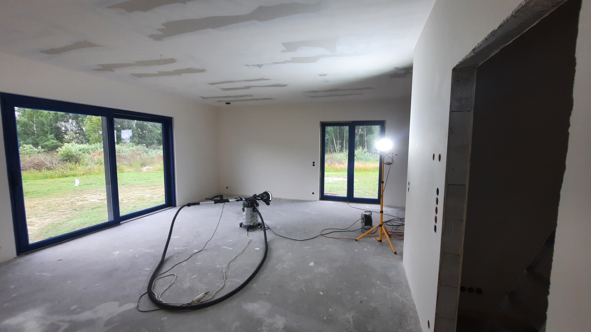 Wnętrze w trakcie prac wykończeniowych: szpachlowanie sufitu, ściany przygotowane do malowania, widoczne narzędzia i lampa oświetleniowa. Duże okna z ciemnymi ramami.