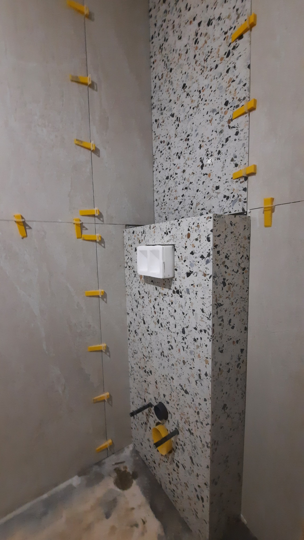 Ściana łazienki w trakcie wykańczania z użyciem płytek w stylu lastryko, z żółtymi klipsami poziomującymi i widoczną instalacją podtynkową.